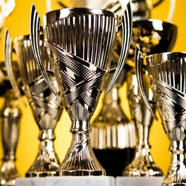 A collection of shiny silver trophies with intricate designs, featuring two handles each and mounted on marble bases, are displayed closely together. The background is a solid yellow color, emphasizing the polished surfaces and reflective qualities of these prestigious awards from top trophy suppliers.