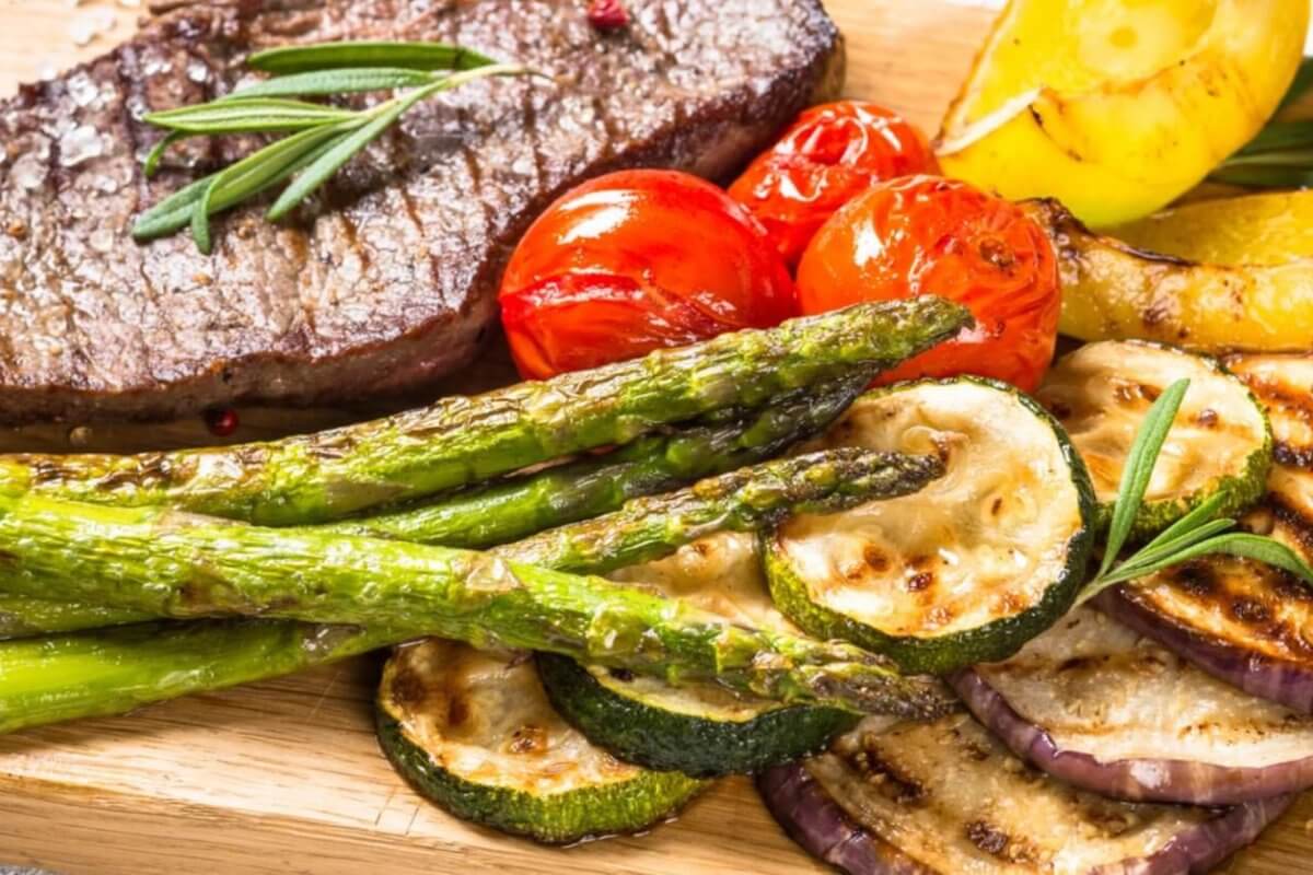 A grilled beef steak garnished with rosemary is served on a wooden board alongside grilled vegetables, including asparagus, zucchini slices, cherry tomatoes, eggplant slices, and bell pepper pieces. This fresh and appetizing meal embodies rustic elegance. Try it at one of the best restaurants in Vertis North.