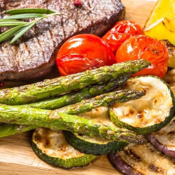 A grilled beef steak garnished with rosemary is served on a wooden board alongside grilled vegetables, including asparagus, zucchini slices, cherry tomatoes, eggplant slices, and bell pepper pieces. This fresh and appetizing meal embodies rustic elegance. Try it at one of the best restaurants in Vertis North.