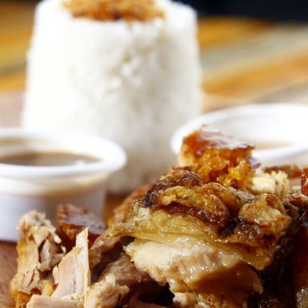 A wooden plate featuring a serving of lechon with crispy skin is shown in the foreground. Two small cups of dipping sauce are placed beside the pork. In the background, a mound of white rice topped with seasoning is visible, presenting a delightful foodie adventure at one of the must-try restaurants in Ayala Center Cebu.
