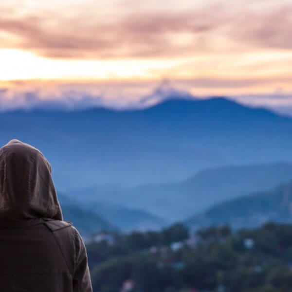 A person wearing a hooded jacket stands with their back to the camera, looking out over a vast, mountainous landscape at sunrise or sunset—a serene moment often experienced during a Baguio Tour. The sky is filled with soft clouds and warm light, casting a serene glow over the rolling hills and valleys below. A second person is partially visible.