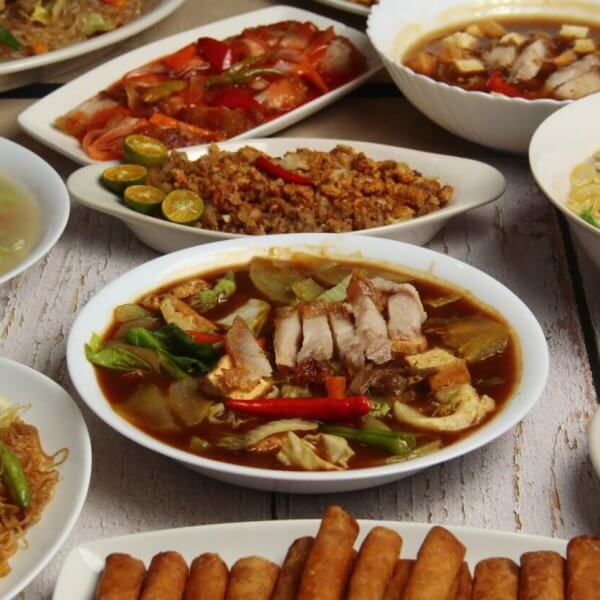 A spread of various Filipino dishes on white plates at one of Banawe, Quezon City's best restaurants. Dishes include bowls of noodle soup with vegetables and meat, a side of spring rolls, stir-fried noodles, grilled meat, and sliced vegetables. The dishes are richly colored with greens, reds, and browns on a white wooden table.