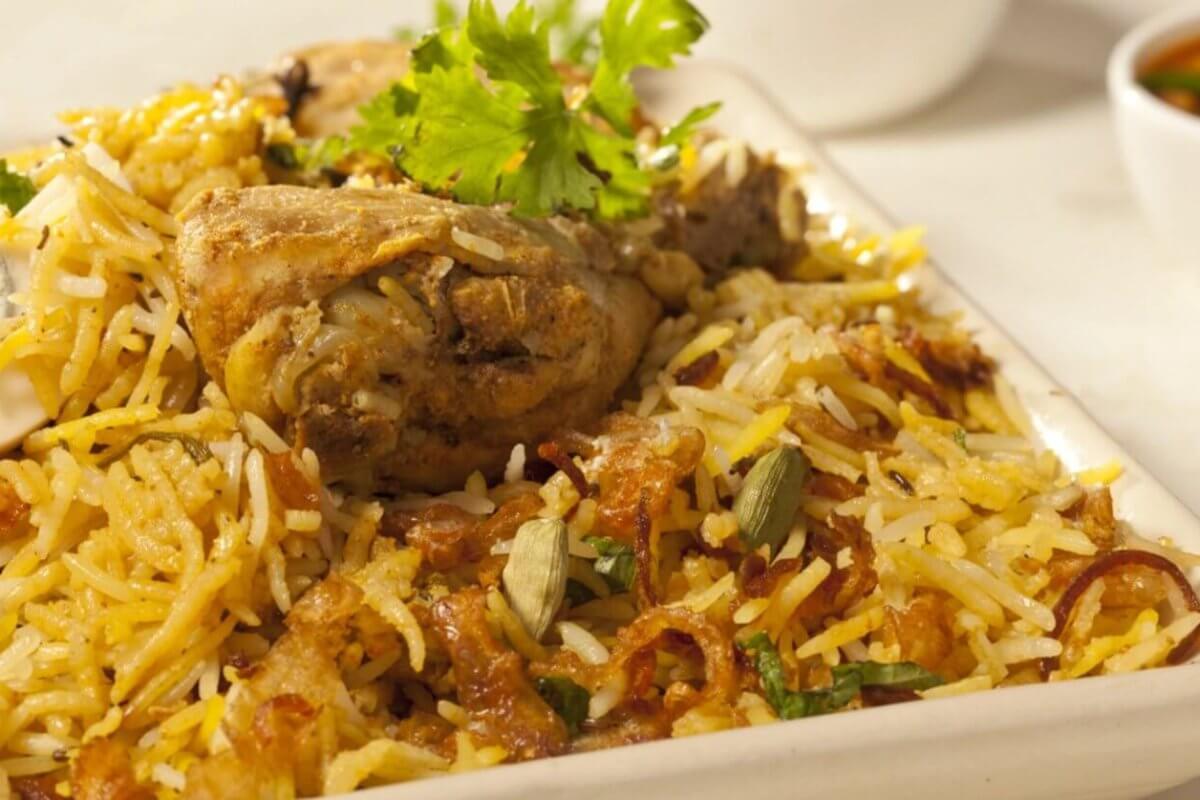 A close-up of a plate of biryani chicken featuring seasoned rice mixed with succulent pieces of chicken, garnished with fresh coriander leaves. This culinary experience is accompanied by a small bowl of curry sauce, slightly blurred in the background. The biryani is colorful, with shades of yellow and brown.