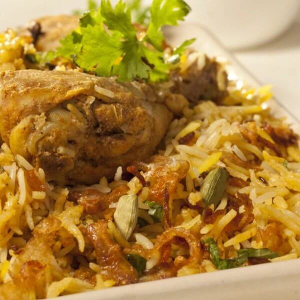 A close-up of a plate of biryani chicken featuring seasoned rice mixed with succulent pieces of chicken, garnished with fresh coriander leaves. This culinary experience is accompanied by a small bowl of curry sauce, slightly blurred in the background. The biryani is colorful, with shades of yellow and brown.