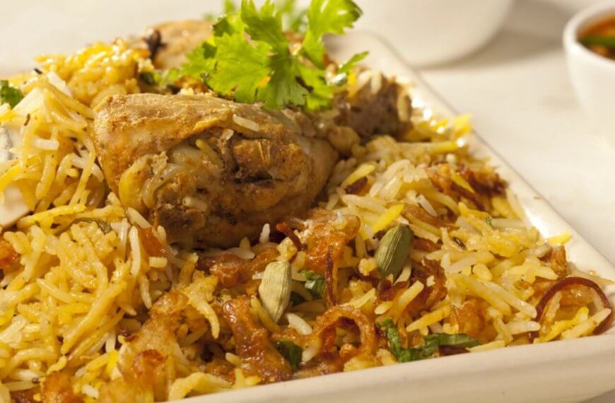 A close-up of a plate of biryani chicken featuring seasoned rice mixed with succulent pieces of chicken, garnished with fresh coriander leaves. This culinary experience is accompanied by a small bowl of curry sauce, slightly blurred in the background. The biryani is colorful, with shades of yellow and brown.