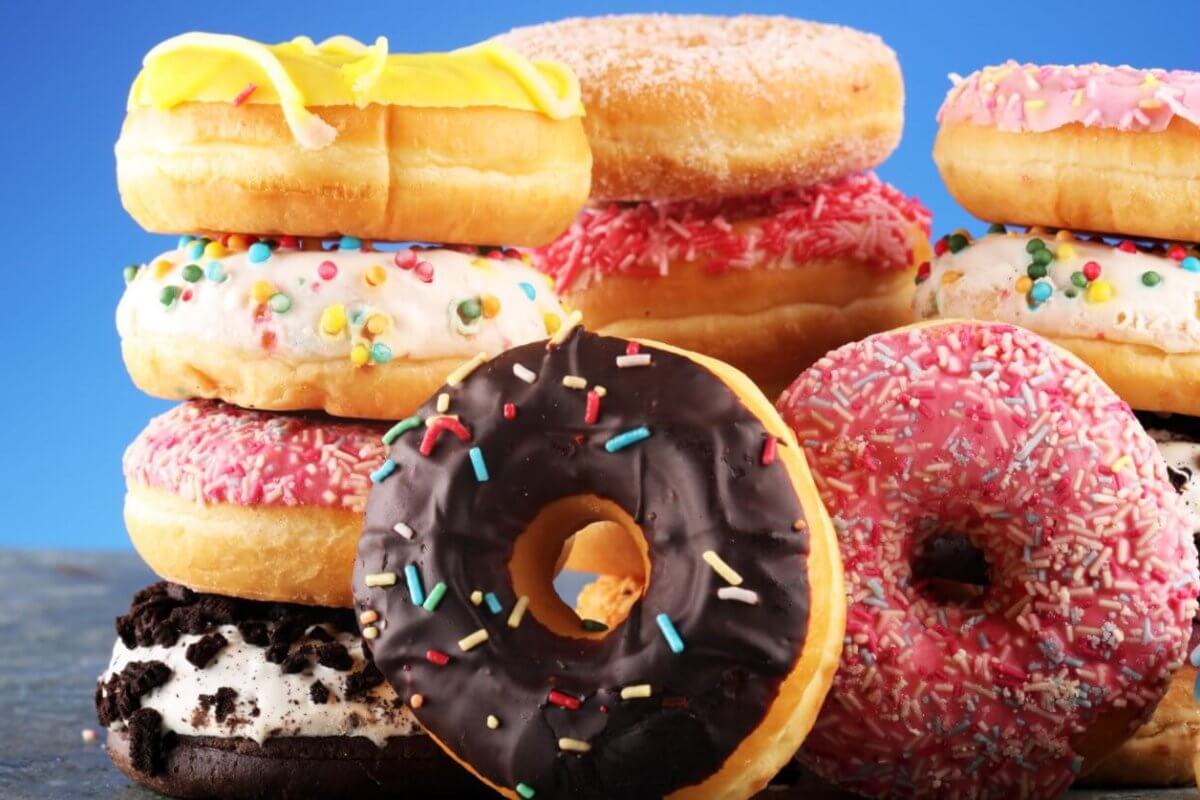 A vibrant assortment of donuts from the best donut shops in Metro Manila is stacked against a blue background. The donuts are topped with various icings and colorful sprinkles, including chocolate, pink, yellow, and white glazes. Some feature additional decorations like cookies and candy bits, making the scene playful and appetizing.