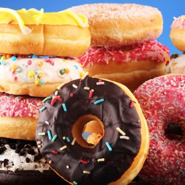 A vibrant assortment of donuts from the best donut shops in Metro Manila is stacked against a blue background. The donuts are topped with various icings and colorful sprinkles, including chocolate, pink, yellow, and white glazes. Some feature additional decorations like cookies and candy bits, making the scene playful and appetizing.