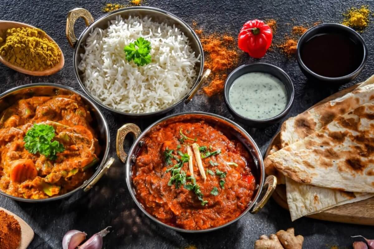 A colorful spread of Indian cuisine on a dark surface featuring bowls of white rice, chicken and vegetable curry, and a rich red curry garnished with cilantro and ginger. Accompanied by naan bread, mint yogurt sauce, and spices like curry powder, turmeric, and paprika—authentic flavors reminiscent of top Indian restaurants in Metro Manila.