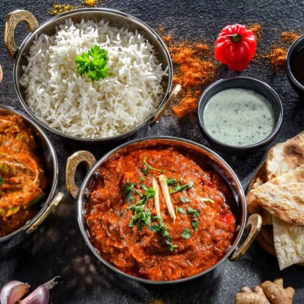 A colorful spread of Indian cuisine on a dark surface featuring bowls of white rice, chicken and vegetable curry, and a rich red curry garnished with cilantro and ginger. Accompanied by naan bread, mint yogurt sauce, and spices like curry powder, turmeric, and paprika—authentic flavors reminiscent of top Indian restaurants in Metro Manila.