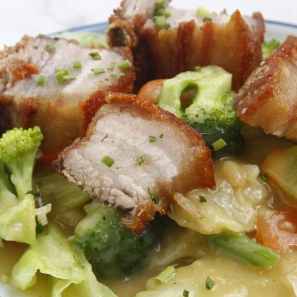 A bowl of crispy pork belly pieces with a golden-brown exterior, served on a bed of vegetables including broccoli, carrots, and cabbage, all coated in a creamy, savory sauce. This dish from one of Nuvali's best restaurants is garnished with finely chopped chives, adding a touch of green.