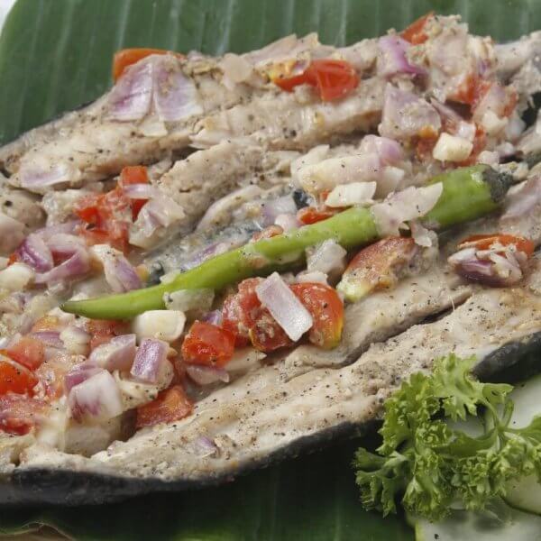 A grilled Bangus split open and laid flat on a green leaf, topped with chopped tomatoes, onions, and garlic, and garnished with a green chili pepper and a small bunch of parsley. Next to the fish are cucumber slices, all arranged on a woven mat—a classic dish you might find at 44 Must-Try Restaurants in Market! Market!.