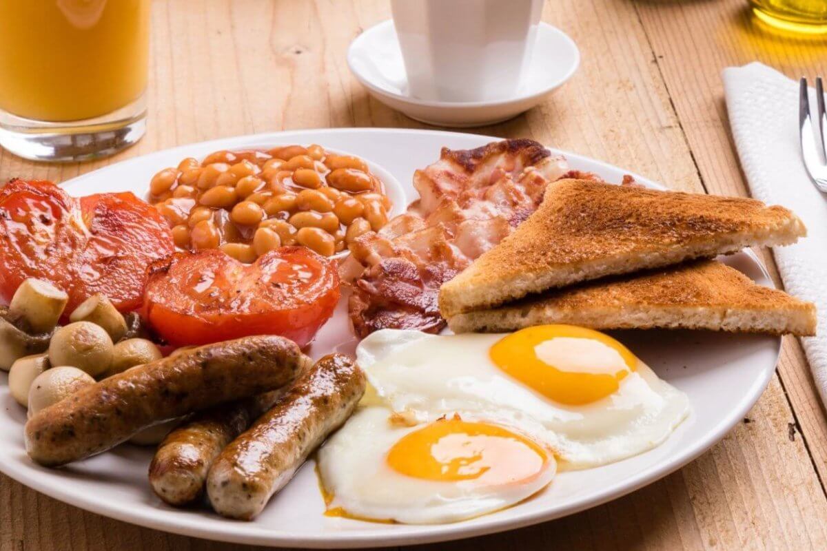 A plate of English breakfast featuring two sunny-side-up eggs, grilled tomatoes, baked beans, crispy bacon, sausages, grilled mushrooms, and two slices of toasted bread. Beside the plate are a fork and knife on a napkin, a glass of orange juice, and a white cup on a wooden table in one of BGC’s best breakfast restaurants.