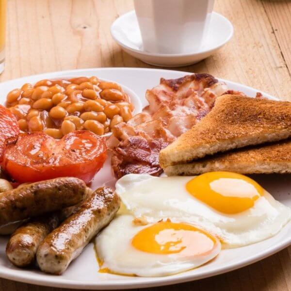 A plate of English breakfast featuring two sunny-side-up eggs, grilled tomatoes, baked beans, crispy bacon, sausages, grilled mushrooms, and two slices of toasted bread. Beside the plate are a fork and knife on a napkin, a glass of orange juice, and a white cup on a wooden table in one of BGC’s best breakfast restaurants.