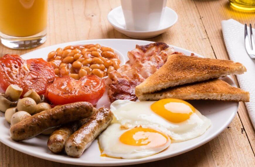 A plate of English breakfast featuring two sunny-side-up eggs, grilled tomatoes, baked beans, crispy bacon, sausages, grilled mushrooms, and two slices of toasted bread. Beside the plate are a fork and knife on a napkin, a glass of orange juice, and a white cup on a wooden table in one of BGC’s best breakfast restaurants.