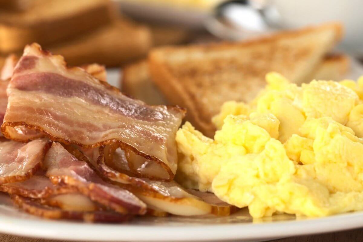 A breakfast plate from one of Marikina's top breakfast restaurants features fluffy scrambled eggs, several strips of crispy bacon with visible fatty edges, and golden brown toast with grill marks.