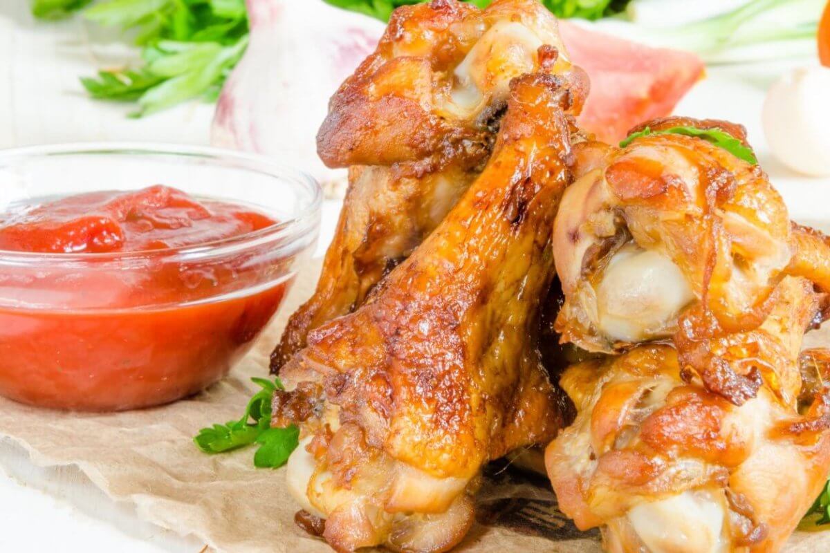 A close-up of golden-brown, crispy Korean chicken wings stacked on a rustic surface, garnished with fresh parsley. A small, clear bowl of red ketchup is positioned to the left. In the blurred background, there are assorted vegetables, including green herbs and garlic cloves—a scene reminiscent of Metro Manila's popular beer spots.