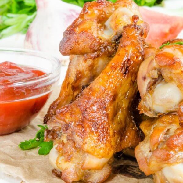 A close-up of golden-brown, crispy Korean chicken wings stacked on a rustic surface, garnished with fresh parsley. A small, clear bowl of red ketchup is positioned to the left. In the blurred background, there are assorted vegetables, including green herbs and garlic cloves—a scene reminiscent of Metro Manila's popular beer spots.