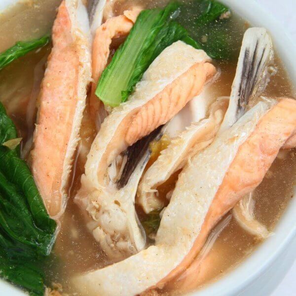 A bowl of clear soup contains pieces of fish, vibrant green leafy vegetables, and orange carrot slices. The fish appears to be salmon, with its distinctive pink color. The broth is light and clear, beautifully presenting a sulit dining experience that could rival the 40 best restaurants in Circuit Manila.