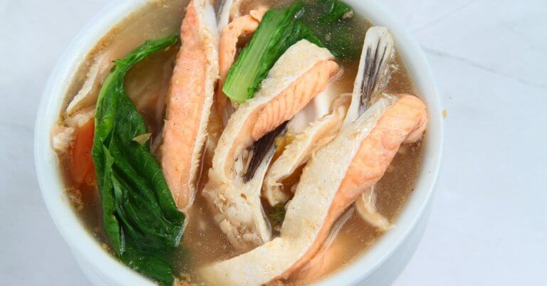 A bowl of clear soup contains pieces of fish, vibrant green leafy vegetables, and orange carrot slices. The fish appears to be salmon, with its distinctive pink color. The broth is light and clear, beautifully presenting a sulit dining experience that could rival the 40 best restaurants in Circuit Manila.