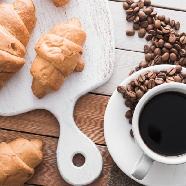 A white cup filled with black coffee from Sulit Coffee sits on a saucer with coffee beans scattered around it on a rustic wooden table. Next to the coffee, there is a wooden cutting board with golden brown croissants placed on it, and more croissants are scattered around the table, evoking the charm of a Metro Manila cafe bakery.
