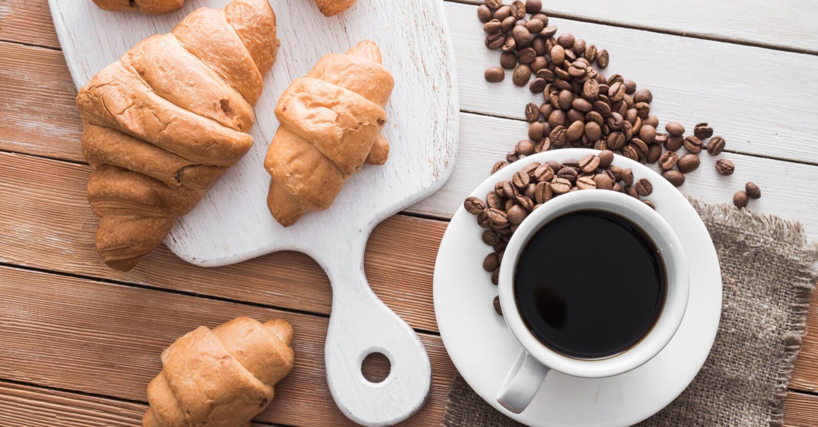A white cup filled with black coffee from Sulit Coffee sits on a saucer with coffee beans scattered around it on a rustic wooden table. Next to the coffee, there is a wooden cutting board with golden brown croissants placed on it, and more croissants are scattered around the table, evoking the charm of a Metro Manila cafe bakery.
