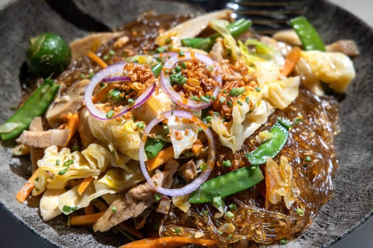 A close-up of a plate of Filipino pancit, featuring glass noodles mixed with sautéed vegetables like cabbage, carrot slices, and snow peas, topped with thinly sliced red onions, fried garlic bits, and chopped green onions. Often found in the best restaurants around Cubao, a green calamansi is seen on the side for garnish.