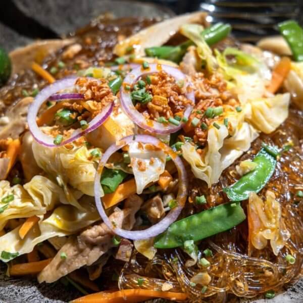 A close-up of a plate of Filipino pancit, featuring glass noodles mixed with sautéed vegetables like cabbage, carrot slices, and snow peas, topped with thinly sliced red onions, fried garlic bits, and chopped green onions. Often found in the best restaurants around Cubao, a green calamansi is seen on the side for garnish.