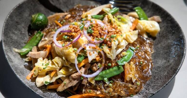 A close-up of a plate of Filipino pancit, featuring glass noodles mixed with sautéed vegetables like cabbage, carrot slices, and snow peas, topped with thinly sliced red onions, fried garlic bits, and chopped green onions. Often found in the best restaurants around Cubao, a green calamansi is seen on the side for garnish.