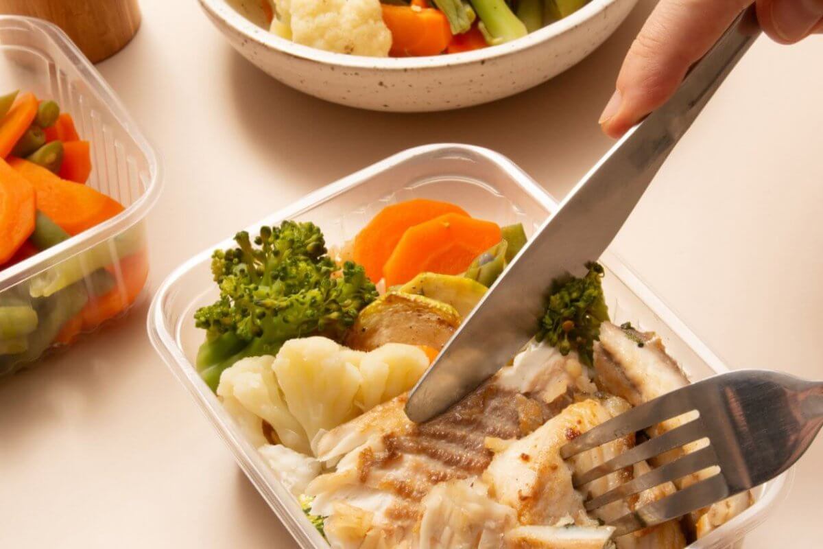 A person using a knife and fork to cut pieces of grilled fish in a plastic container. The container also holds cooked broccoli, cauliflower, and carrots. Additional containers filled with more steamed mixed vegetables and a white bowl are in the background, ideal for healthy meal plans from diet delivery services.