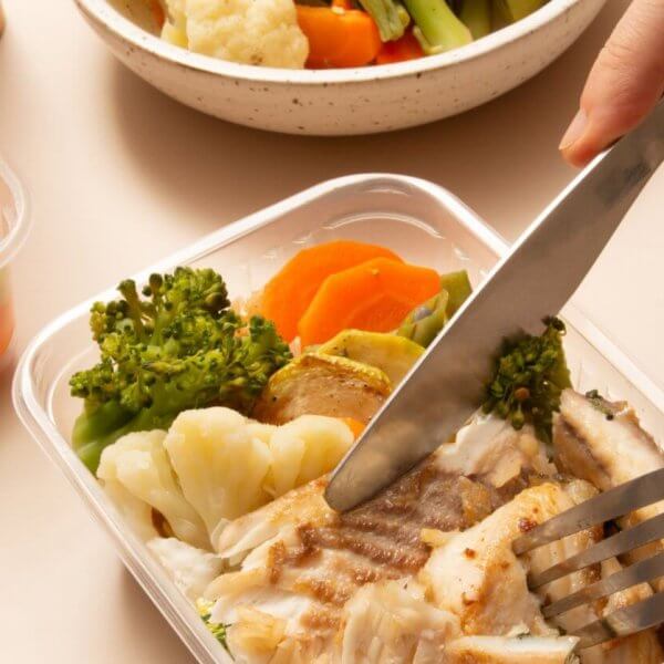 A person using a knife and fork to cut pieces of grilled fish in a plastic container. The container also holds cooked broccoli, cauliflower, and carrots. Additional containers filled with more steamed mixed vegetables and a white bowl are in the background, ideal for healthy meal plans from diet delivery services.