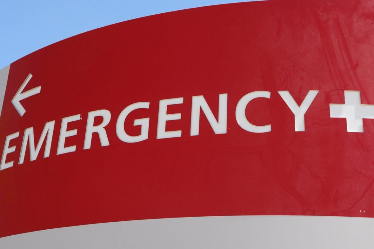 A red and white hospital emergency sign is displayed. The word "EMERGENCY" and a plus symbol are prominently written in white uppercase letters. A white arrow points left, directing towards the Emergency Rescue Services area. The background includes a blue sky and a hint of green foliage. 