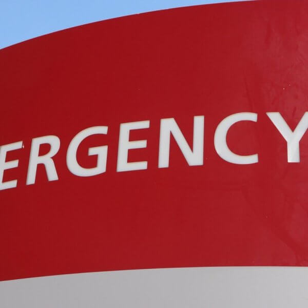 A red and white hospital emergency sign is displayed. The word "EMERGENCY" and a plus symbol are prominently written in white uppercase letters. A white arrow points left, directing towards the Emergency Rescue Services area. The background includes a blue sky and a hint of green foliage.
