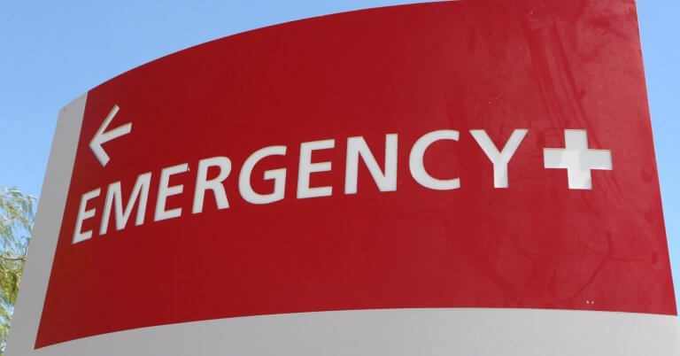 A red and white hospital emergency sign is displayed. The word "EMERGENCY" and a plus symbol are prominently written in white uppercase letters. A white arrow points left, directing towards the Emergency Rescue Services area. The background includes a blue sky and a hint of green foliage.
