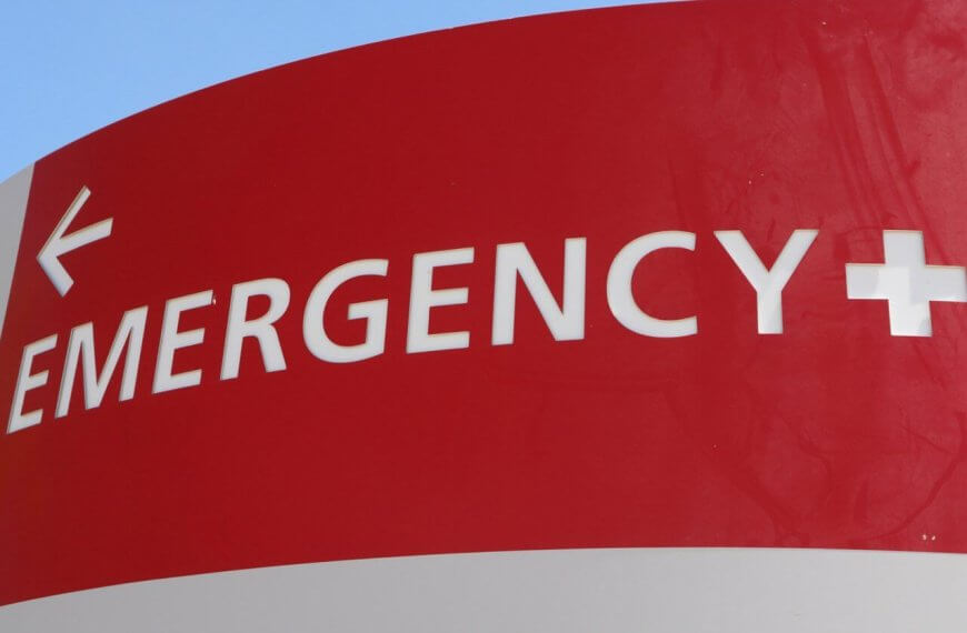 A red and white hospital emergency sign is displayed. The word "EMERGENCY" and a plus symbol are prominently written in white uppercase letters. A white arrow points left, directing towards the Emergency Rescue Services area. The background includes a blue sky and a hint of green foliage.
