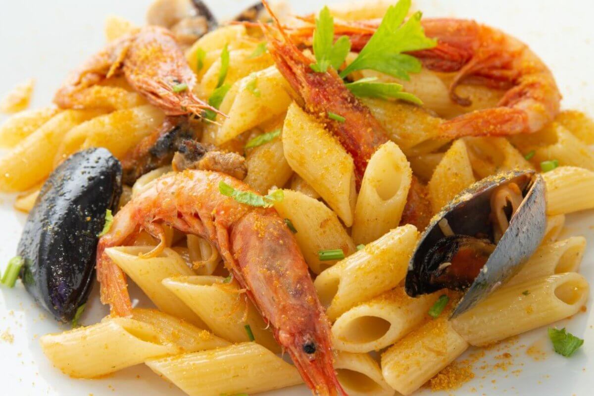 A close-up of a seafood pasta dish featuring penne pasta, large shrimp, and mussels. Garnished with sprigs of fresh parsley and a light dusting of seasoning, the dish is served on a square white plate at one of the best restaurants in Evia Lifestyle Center. The vibrant colors contrast with the creamy pasta.