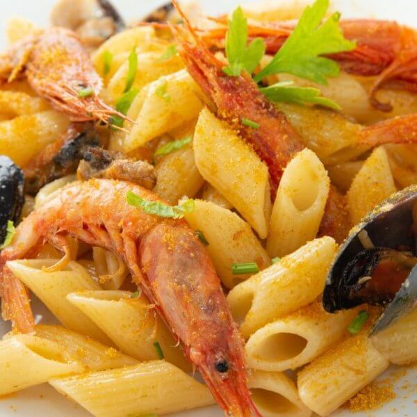 A close-up of a seafood pasta dish featuring penne pasta, large shrimp, and mussels. Garnished with sprigs of fresh parsley and a light dusting of seasoning, the dish is served on a square white plate at one of the best restaurants in Evia Lifestyle Center. The vibrant colors contrast with the creamy pasta.