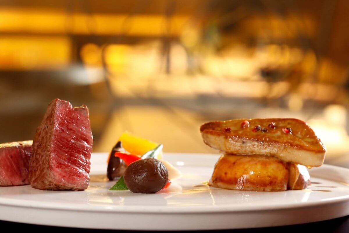 A gourmet dish on a white plate features a neatly cut piece of medium-rare beef, an artistic arrangement of colorful vegetables, a dollop of dark sauce, and seared foie gras atop a round base. This fine dining experience in Metro Manila is beautifully highlighted against a softly blurred background.