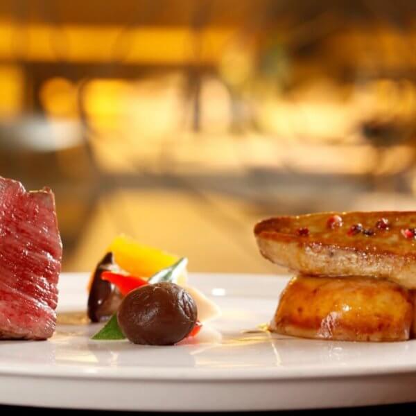 A gourmet dish on a white plate features a neatly cut piece of medium-rare beef, an artistic arrangement of colorful vegetables, a dollop of dark sauce, and seared foie gras atop a round base. This fine dining experience in Metro Manila is beautifully highlighted against a softly blurred background.