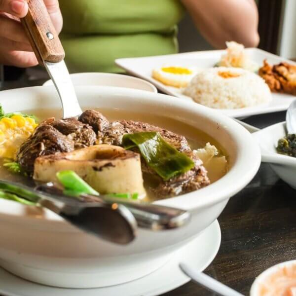 A person in a green shirt is cutting meat in a bowl of soup with a knife and fork. The table is set with iced tea, a plate of rice with fried food, and a dish of greens, highlighting the Best Restaurants. Small plates around feature a variety of foods, making it the Ultimate Glorietta Experience.