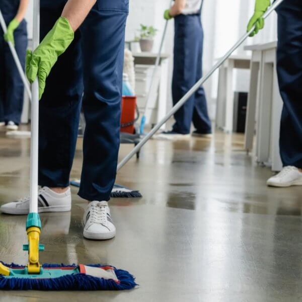 Cleaning people in blue uniforms and green gloves are mopping the floor in an office setting, representing Sulit Clean's Home Cleaning Services. The focus is on the person in the foreground, visible from the waist down, using a flat mop. White desks, office chairs, and trash cans fill out the Metro Manila background.