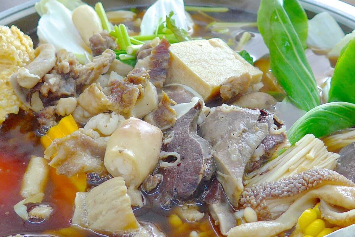 An assortment of ingredients simmering in a hot pot filled with broth. Visible items include various cuts of meat, tofu, mushrooms, leafy greens, pieces of corn, and a chunk of tomato. The ingredients float in a rich brown broth, creating a hearty, communal meal—perfect for those looking to spice up their taste buds at the best hotpot restaurants in Metro Manila.