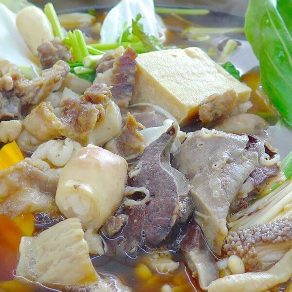 An assortment of ingredients simmering in a hot pot filled with broth. Visible items include various cuts of meat, tofu, mushrooms, leafy greens, pieces of corn, and a chunk of tomato. The ingredients float in a rich brown broth, creating a hearty, communal meal—perfect for those looking to spice up their taste buds at the best hotpot restaurants in Metro Manila.