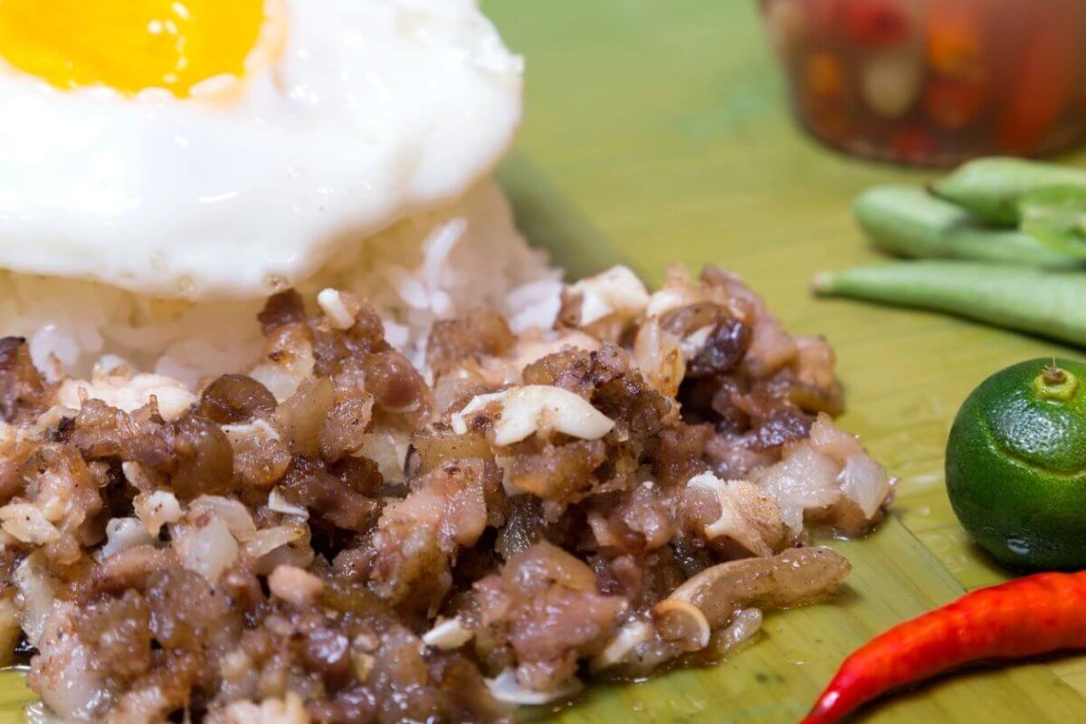 A close-up of traditional Filipino food featuring a serving of garlic rice topped with a sunny-side-up egg. Adjacent to the rice is a portion of sizzling chopped meat, likely pork. Garnishing the plate are a whole calamansi, a red chili pepper, and a few green beans. A condiment jar is blurred in the background.