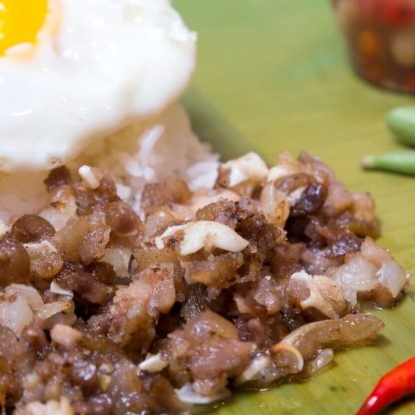 A close-up of traditional Filipino food featuring a serving of garlic rice topped with a sunny-side-up egg. Adjacent to the rice is a portion of sizzling chopped meat, likely pork. Garnishing the plate are a whole calamansi, a red chili pepper, and a few green beans. A condiment jar is blurred in the background.