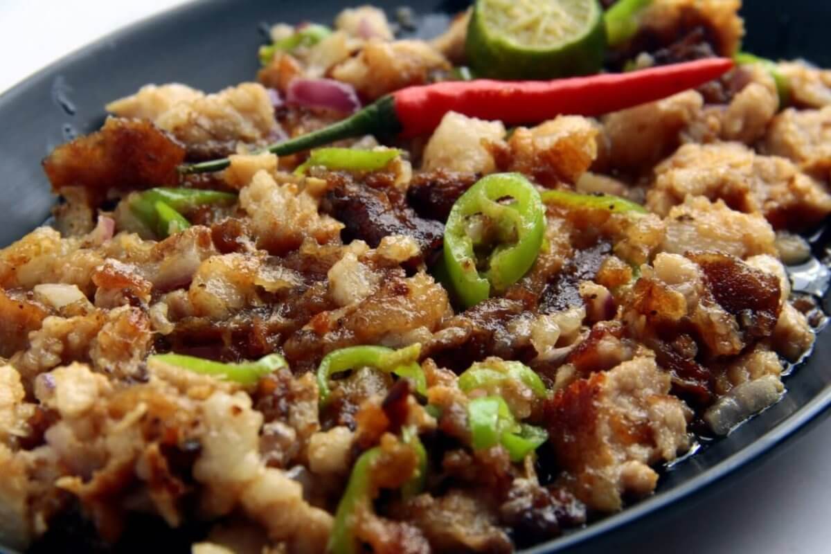 A black oval plate holds a serving of sisig, a popular Filipino dish. This Food Lover's Guide favorite from Laguna consists of chopped pork, onions, and green chili slices topped with a whole red chili. A sliced calamansi lime and crispy bits of pork adorn the top, with visible oil glistening on the surface.