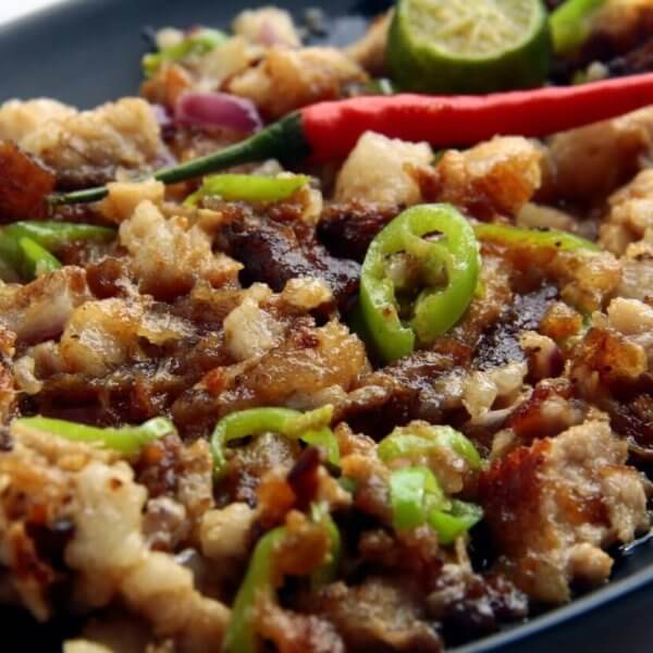 A black oval plate holds a serving of sisig, a popular Filipino dish. This Food Lover's Guide favorite from Laguna consists of chopped pork, onions, and green chili slices topped with a whole red chili. A sliced calamansi lime and crispy bits of pork adorn the top, with visible oil glistening on the surface.