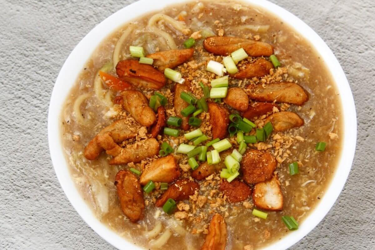 A top-down view of a hearty bowl of noodle soup garnished with chunks of fried chicken, sliced green onions, and bits of crunchy fried garlic. The soup's rich broth is visible, with noodles submerged beneath the toppings. This delectable dish could rival the best Lomi places in Batangas. The bowl rests on a textured gray surface.