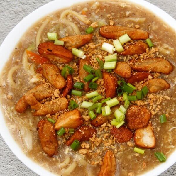 A top-down view of a hearty bowl of noodle soup garnished with chunks of fried chicken, sliced green onions, and bits of crunchy fried garlic. The soup's rich broth is visible, with noodles submerged beneath the toppings. This delectable dish could rival the best Lomi places in Batangas. The bowl rests on a textured gray surface.