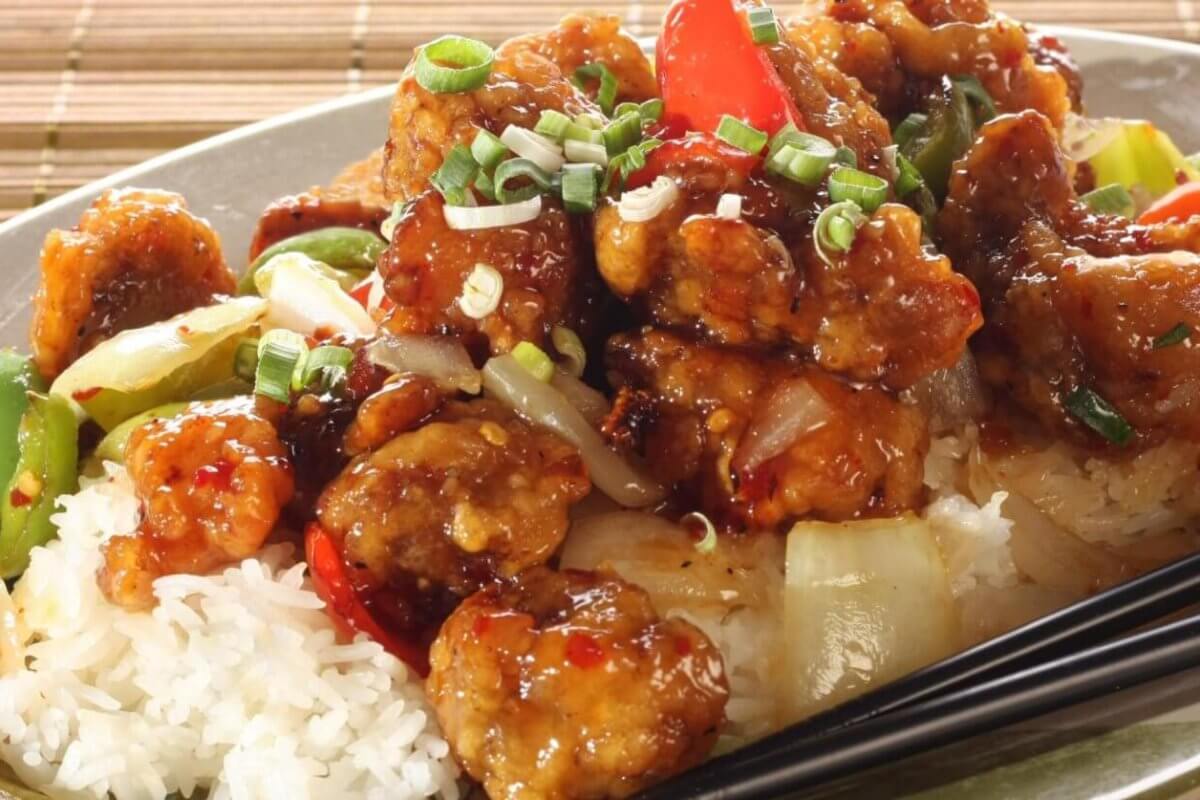 A plate filled with sweet and sour chicken served over white rice is garnished with chopped green onions. The dish includes chunks of chicken coated in a glossy sauce with red and green bell peppers and onions. Black chopsticks rest on the side of the plate, which sits on a wooden surface at Lucky Chinatown.
