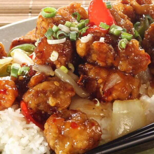 A plate filled with sweet and sour chicken served over white rice is garnished with chopped green onions. The dish includes chunks of chicken coated in a glossy sauce with red and green bell peppers and onions. Black chopsticks rest on the side of the plate, which sits on a wooden surface at Lucky Chinatown.